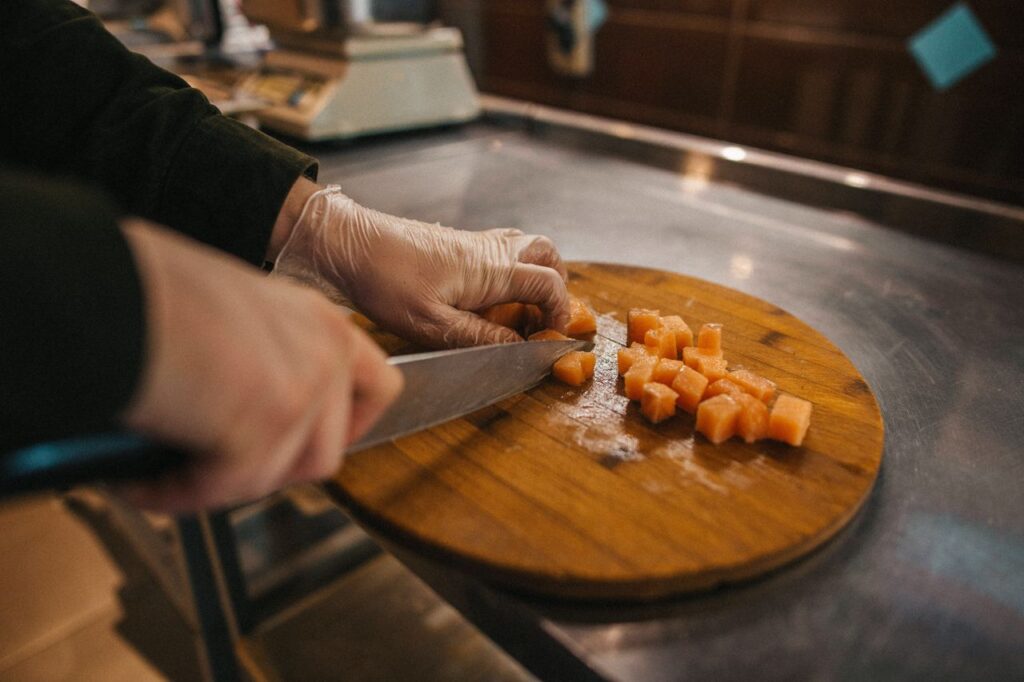 Chef wearing gloves slices fresh salmon on a wooden cutting board in a professional kitchen.