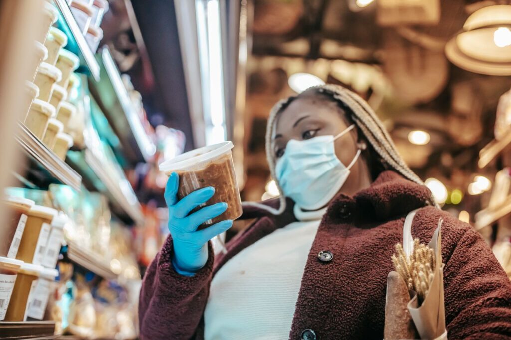 Low angle of African American female in mask and latex gloves considering food while shopping in supermarket during pandemic of coronavirus